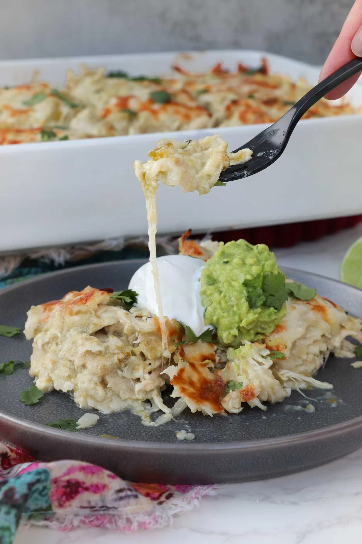 Plate with a single serving of Chicken Enchilada Casserole, topped with sour cream, guacamole, and cilantro. A fork with a bite of casserole is held, raised, above the plate, a string of cheese connecting the bite to the plated casserole serving. The rest of the casserole rests in the background.
