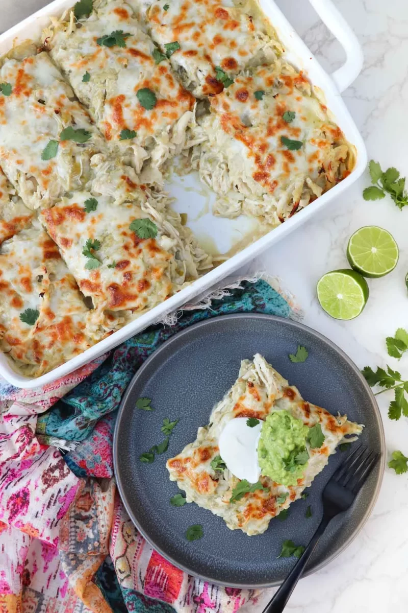 Overhead picture of a plate with a single serving of Chicken Enchilada Casserole in the bottom right corner, with the baking dish with the rest of the casserole in the top left. A halved lime rests near the plate, and a colorful linen sits under the plate.